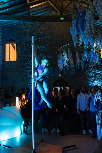 Nathalie effectue une figure de pole dance devant un public lors d’un événement festif en intérieur, sous une lumière bleue et des décorations florales suspendues