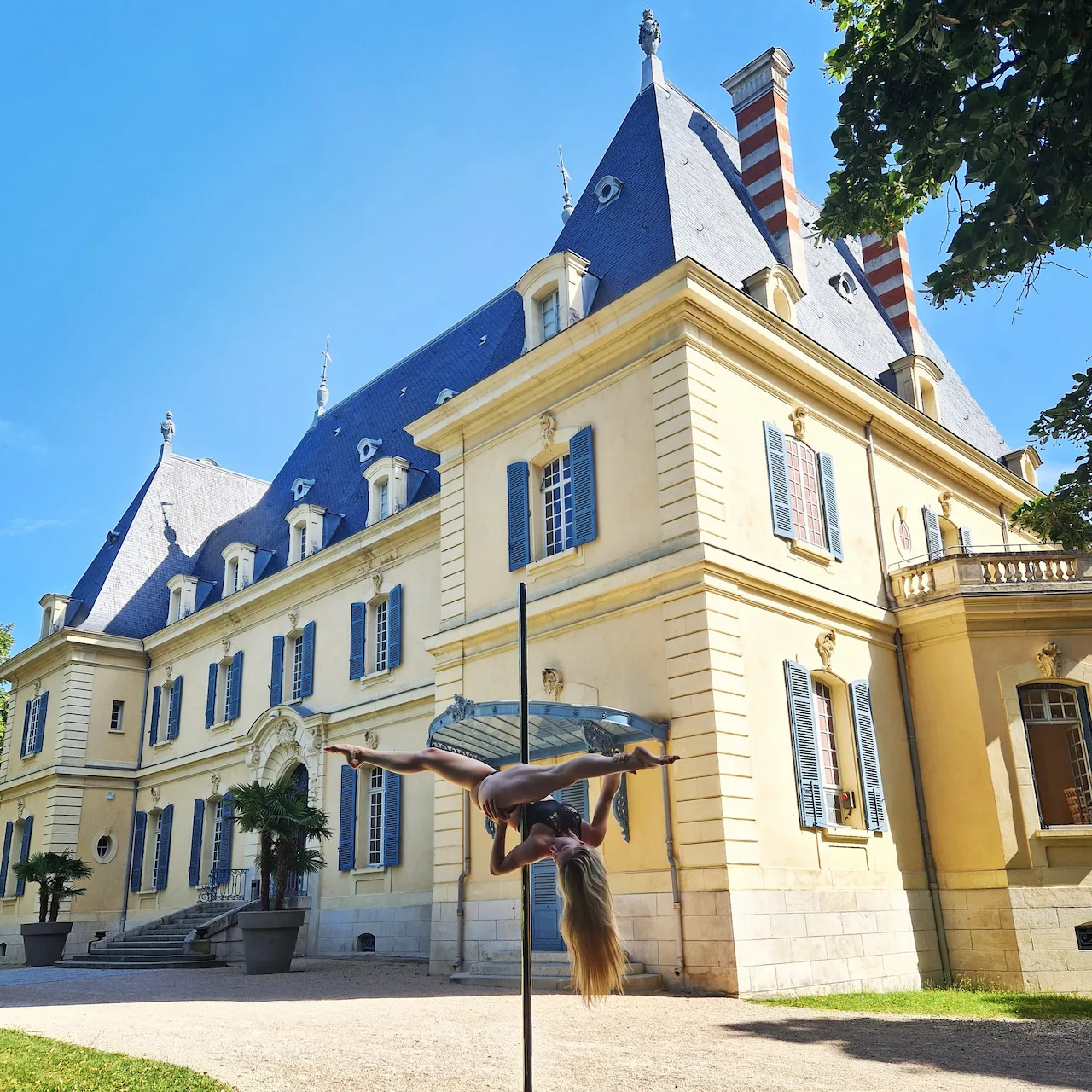 Madame Passions réalisant une figure de pole dance en extérieur, devant un château